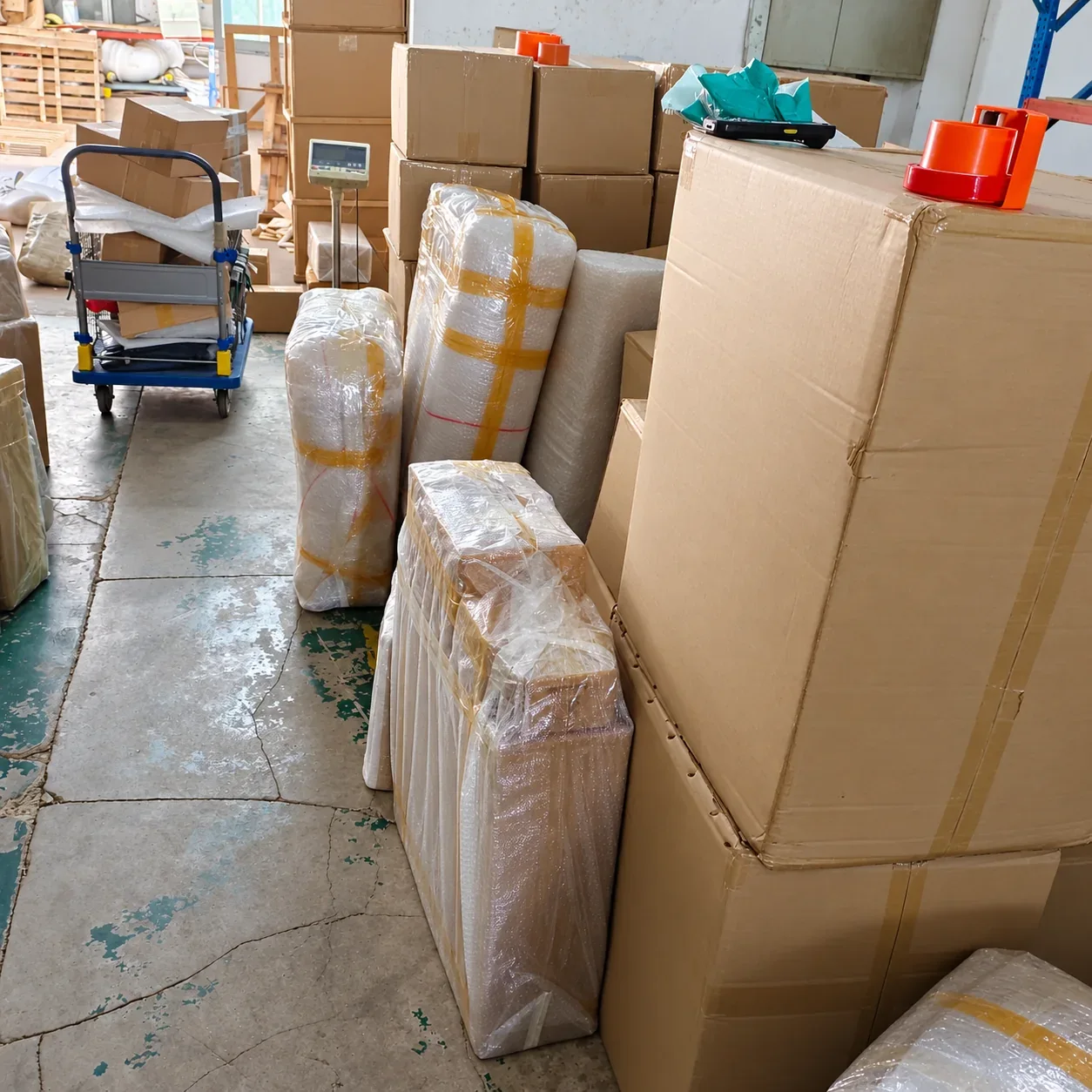 Protective wrapping around long parcels beside a trolley and stacked cartons.