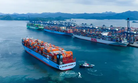 Container ship crossing blue harbour water beside a long row of quay cranes