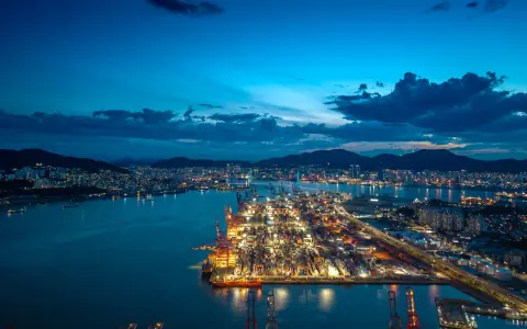 Blue-hour container port, harbour water and city lights beneath dark clouds
