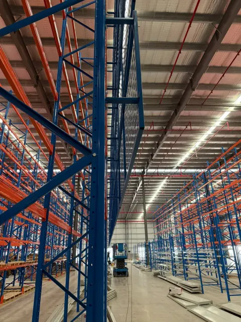 Tall blue and orange storage racks beneath a warehouse roof